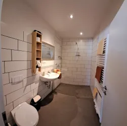 Ein modernes Badezimmer mit weißen Fliesen und einem Waschbecken. Es gibt eine begehbare Dusche, eine Toilette und einen großen Handtuchhalter.