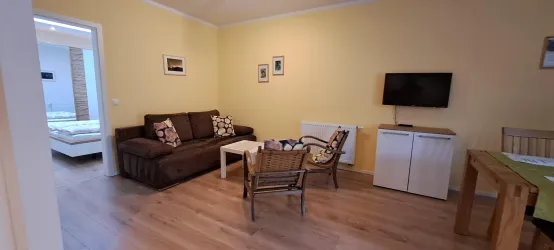 Ein gemütliches Wohnzimmer mit einem braunen Sofa und einem Fernseher an der Wand. Der Raum hat helle Wände und einen Holzboden, mit einem kleinen Tisch und Stühlen.