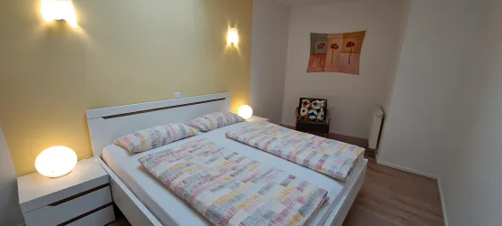 Ein gemütliches Schlafzimmer mit einem großen Doppelbett und hellen Wänden. Es gibt moderne Lampen und ein Kunstwerk an der Wand.