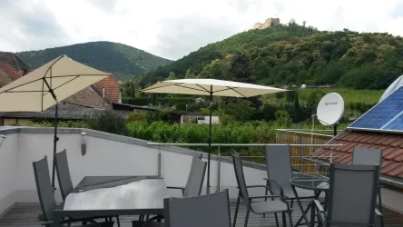 Eine Terrasse mit Tischen und Stühlen, umgeben von grünen Hügeln. Im Hintergrund ist das Hambacher Schloss zu sehen.