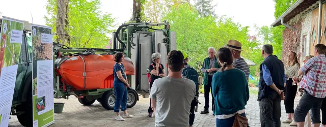 Eine Gruppe von Menschen hört einem Vortrag in einem Garten oder Hof zu. Im Hintergrund steht ein Traktor mit einer großen orangefarbenen Anlage.