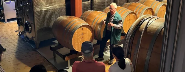 Eine Weinprobe im Keller, in dem Holzfässer stehen. Ein älterer Mann erklärt den Besuchern den Weinherstellungsprozess.