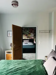 Ein gemütliches Schlafzimmer mit einem grünen Bett und einem offenen Türbogen, der zu einem weiteren Raum führt. Der Raum hat helle Wände und eine dekorative Tapete mit Blumenmuster.