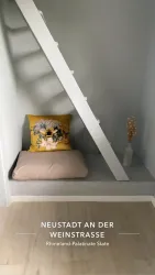 Eine gemütliche Ecke mit einem Kissen und einem dekorativen Kissen auf einer Holzbank. Die Wand ist in hellen Farben gestrichen, und eine Treppe führt nach oben.