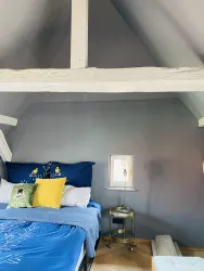 Ein gemütliches Schlafzimmer mit einem blauen Bett und bunten Kissen. Die Wände sind grauweiß und die Holzbeamensicht bietet einen modernen, rustikalen Look.