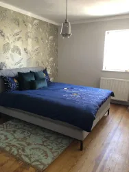 Ein modernes Schlafzimmer mit einem großen Bett und bunten Kissen. Die Wände sind mit einem eleganten Muster dekoriert und ein Fenster lässt viel Licht herein.