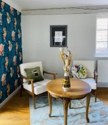 Ein gemütlicher Wohnraum mit zwei Stühlen und einem runden Tisch. Die Wände sind mit einer blumigen Tapete und einem modernen Bild dekoriert.