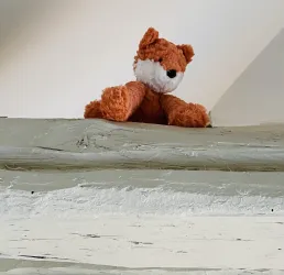 Ein orangefarbener Plüschfuchs sitzt auf einer Holzoberfläche. Der Hintergrund ist hell und neutral.