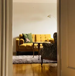 Ein gemütliches Wohnzimmer mit einem gelben Sofa und einem Sessel. Der Teppich sorgt für eine einladende Atmosphäre.