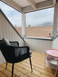 Ein gemütlicher Balkon mit einem schwarzen Stuhl und einem kleinen Tisch. Die Aussicht zeigt Dächer und eine bergige Landschaft im Hintergrund.