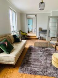 Ein modernes Wohnzimmer mit einem beigen Sofa und einem Glas-Tisch. Der Raum hat Holzböden und große Fenster, die viel Tageslicht hereinlassen.