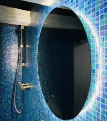 Ein stilvolles Bad mit blauen Mosaikfliesen und einem großen ovalen Spiegel. Der Spiegel hat eine integrierte Beleuchtung, die den Raum erhellt.