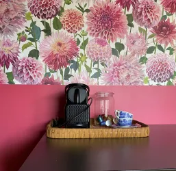 Ein dekorativer Raum mit einer blühenden Blumenwand in Pinktönen. Auf einem Tisch befindet sich eine Kaffeekapselmaschine und eine Tasse.