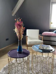Ein modernes Wohnzimmer mit einem gemütlichen Sessel und zwei eleganten Beistelltischen. Auf einem Tisch steht eine Vase mit bunten Trockenblumen.
