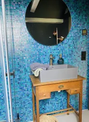 Ein modernes Badezimmer mit blauen Mosaikfliesen. Ein Holzschrank mit einer grauen Waschschüssel und einem runden Spiegel.