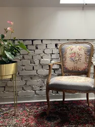 Ein eleganter Stuhl mit floralem Muster steht vor einer Ziegelwand. Daneben befindet sich ein Topf mit einer Pflanze auf einem goldenen Gestell.