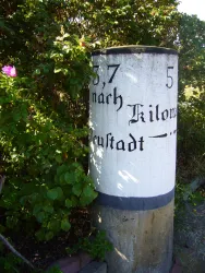 Ein Wegweiser zeigt die Entfernung nach Kiel und Neustadt an. Der Pfeiler ist von Pflanzen umgeben.