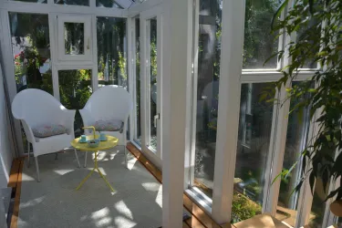 Eine helle Veranda mit zwei weißen Stühlen und einem kleinen gelben Tisch. Viele Pflanzen und Licht durch die Fenster schaffen eine entspannte Atmosphäre.