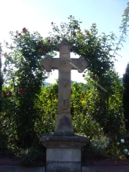 Ein steinernes Kreuz steht umgeben von üppigem Grün und blühenden Rosen. Der Himmel ist klar und blau, was eine ruhige Atmosphäre schafft.