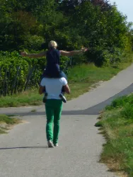 Ein Erwachsener trägt ein Kind auf den Schultern und geht einen Weg entlang. Umgeben von Bäumen und grüner Landschaft strahlt die Szene Freude aus.
