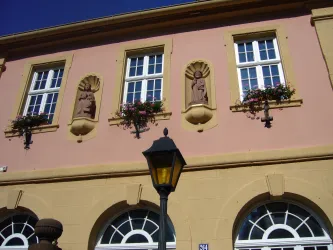 Eine historische Fassade mit pinkem Putz und dekorativen Skulpturen. Unter den Fenstern hängen Blumenkästen und eine Straßenlaterne steht im Vordergrund.