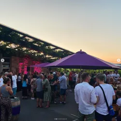 Eine lebhafte Außenveranstaltung mit vielen Menschen unter großen Sonnenschirmen. Im Hintergrund ist ein schöner Sonnenuntergang zu sehen.