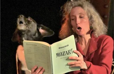 Eine Frau singt laut und hält ein Notenbuch mit dem Titel "Mozart". Neben ihr steht ein Hund, der ebenfalls den Kopf in die Höhe richtet.