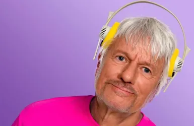 Ein älterer Mann mit grauen Haaren trägt gelbe Kopfhörer. Er hat ein freundliches Lächeln und trägt ein pinkes T-Shirt.