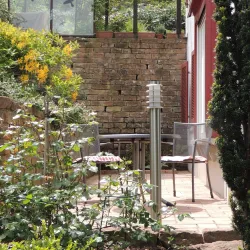 Eine gemütliche Terrasse mit einem Tisch und Stühlen, umgeben von grünen Pflanzen und gelben Blumen. Im Hintergrund sieht man eine Bankmauer aus Steinen.