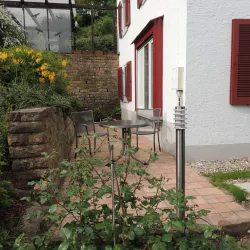 Ein gemütlicher Patio neben einem Haus mit einem Tisch und zwei Stühlen. Umgeben von grünen Pflanzen und einem Stein.