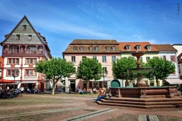 Ein schöner Platz mit bunten Gebäuden und Bäumen. In der Mitte befindet sich ein Brunnen, umgeben von Menschen, die die Sonne genießen.