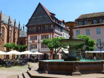 Ein malerischer Platz mit einem Brunnen und traditionellen Gebäuden. In der Umgebung stehen Bäume und Tische mit Sonnenschirmen.