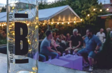 Eine glasflasche mit Lichtern steht im Vordergrund. Im Hintergrund sieht man eine gesellige Menschenmenge bei einer Veranstaltung im Freien.