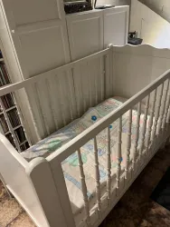 Ein weißes Babybett mit einem bunten Bettlaken. Es steht in einem Raum mit Schränken und Holzfußboden.