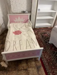 Ein kleines Kinderbett mit einer rosa Krone und einer Bettdecke, die das Wort "DREAM" zeigt. Der Boden ist mit einem Teppich aus bunten Mustern bedeckt.