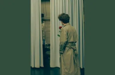 Ein Mann in einem langen Mantel steht mit einer roten Rose in der Hand vor einem Vorhang. Er blickt nachdenklich in den Raum.