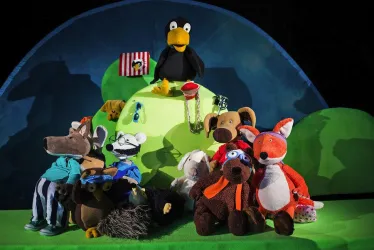 Eine bunte Bühne mit verschiedenen Plüschfiguren, darunter ein Waschbär, ein Hund und ein Fuchs. Im Hintergrund steht ein gelber Vogel und es gibt eine grüne Landschaft.