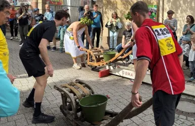 Zwei Teilnehmer bei einem Wettrennen mit Schubkarren auf einer belebten Straße. Zuschauer beobachten gespannt das Geschehen.