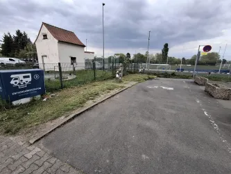 Ein Parkplatz mit einem Gehweg und einem kleinen Gebäude im Hintergrund. Der Himmel ist bewölkt und die Umgebung scheint ruhig zu sein.
