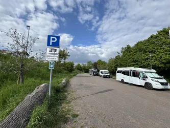 Ein Parkplatz mit einem Parkplatzschild und zwei Fahrzeugen auf einer gravel Straße. Im Hintergrund sind Bäume und ein blauer Himmel mit Wolken zu sehen.