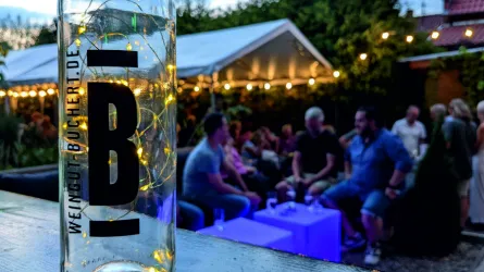 Ein festlicher Außenbereich mit Gästen, die sich unter Lichtern entspannen. Im Vordergrund steht eine Flasche mit dem Schriftzug „Weingut Buchert“.