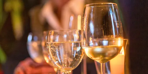 Gläser mit Champagner stehen auf einem Tisch. Im Hintergrund sind Kerzenlicht und verschwommene Personen zu sehen.