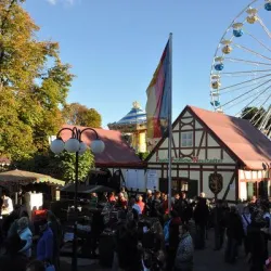 Ein belebter Jahrmarkt mit vielen Menschen und festlicher Atmosphäre. Im Hintergrund sind ein großes Riesenrad und bunte Stände zu sehen.