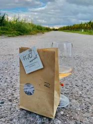 Ein Glas Wein steht auf einem Kiesweg, daneben eine braune Papierverpackung. Im Hintergrund sind Weinreben und ein bewölkter Himmel zu sehen.