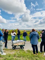 Eine Gruppe von Menschen steht in einem Weinberg unter einem bewölkten Himmel. Im Vordergrund steht ein Wagen mit Materialien.