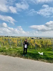 Eine Person steht inmitten von Weinbergen und hält ein Blatt Papier in der Hand. Neben ihr steht ein kleiner Hund und die Wolken sind teilweise am Himmel zu sehen.