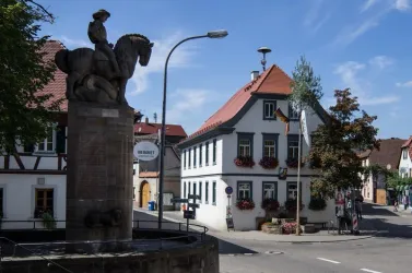 Eine charmante Stadtansicht mit einem Denkmal eines Reiters und einem historischen Gebäude im Hintergrund. Der Himmel ist klar und blau, und die Straßen sind mit Bäumen gesäumt.