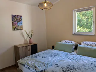 Ein gemütliches Schlafzimmer mit einem Doppelbett und frischem Bettzeug. An der Wand hängt ein farbenfrohes Bild, und das Fenster lässt viel Tageslicht rein.