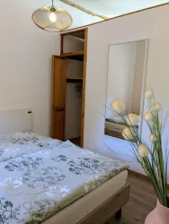 Ein heller, einladender Raum mit einem Bett und einem großen Spiegel. Eine Vase mit dekorativen Blumen sorgt für einen schönen Akzent im Raum.