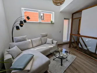 Eine gemütliche Sitzecke mit einem grauen Sofa und einem Glastisch. Das Zimmer ist hell und modern, mit großen Fenstern und Holzdetails.